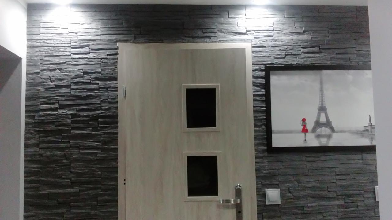 Ściana wykończona dekoracyjną okładziną z szarego kamienia łupkowego, oświetlona od góry, sąsiadująca z drzwiami z dwoma małymi okienkami i obrazem z motywem wieży Eiffla.