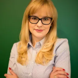 Portret kobiety w okularach, blond włosy, skrzyżowane ręce, na zielonym tle.