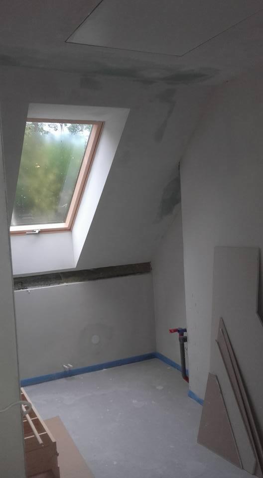 Pokój na poddaszu w trakcie prac wykończeniowych: okno dachowe z widokiem na zieleń, surowe ściany z widocznymi plamami, zabezpieczona taśmą podłoga i rury, płyty gipsowe oparte o ścianę.