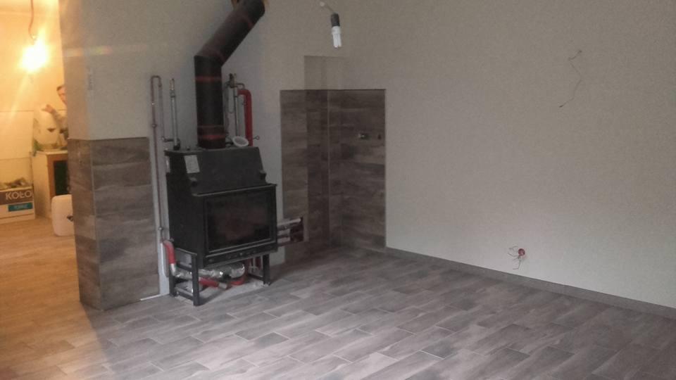 Wnętrze pokoju z kominkiem z czarnym wkładem i rurą dymową, szare podłogi i okładziny ścienne imitujące drewno, widoczne przygotowane przyłącze elektryczne na ścianie.