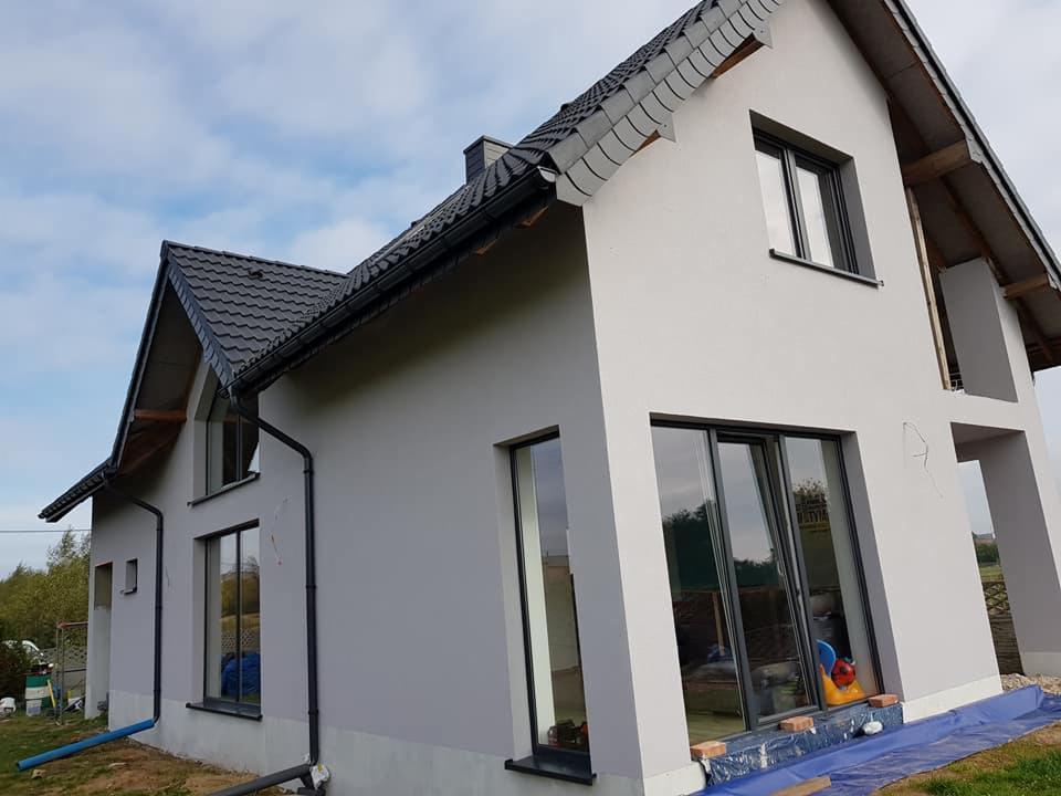 Szary dom jednorodzinny z ciemną dachówką i oknami, widoczne przygotowania do dalszych prac wykończeniowych wokół okien.