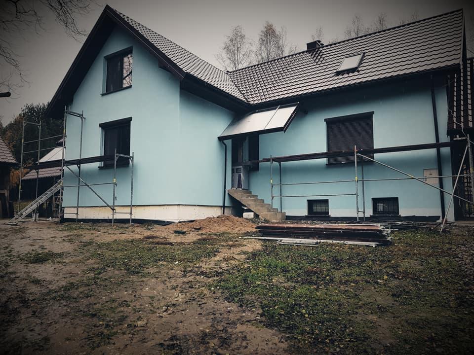 Dom jednorodzinny z niebieską elewacją w trakcie malowania, widoczne rusztowania i świeżo pomalowana podmurówka, teren wokół domu częściowo rozkopany.