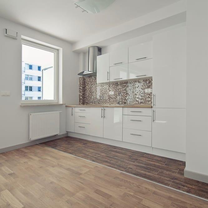 Jasna kuchnia z białymi szafkami, mozaikową ścianą nad blatem i dwoma rodzajami podłogi: jasne panele i ciemne płytki imitujące drewno.