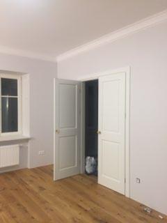 Pomalowane na jasny fiolet pokój z drewnianą podłogą, białymi drzwiami dwuskrzydłowymi i listwami przysufitowymi. Widoczne okno z białymi ramami i grzejnikiem pod nim.