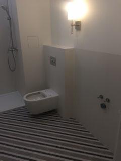 Nowoczesna łazienka z podwieszaną toaletą, szarymi pasami na podłodze i minimalistyczną armaturą. Widoczny natrysk z deszczownicą i oświetlenie ścienne.