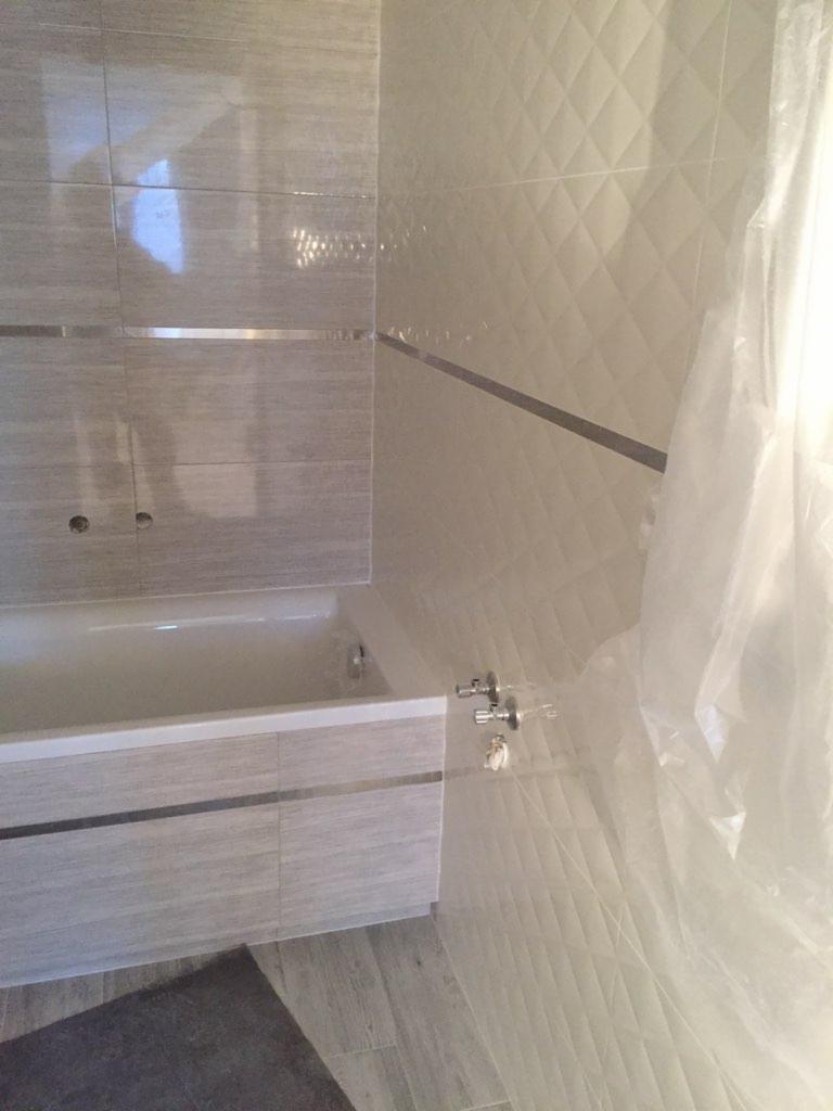 Wykończona łazienka z wanną obudowaną płytkami imitującymi drewno, ściany pokryte płytkami w dwóch wzorach: gładkie w pasy i z wypukłym wzorem rombów, oddzielone metalową listwą.