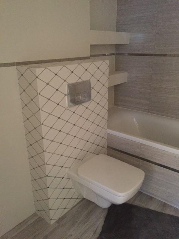 Nowoczesna toaleta podwieszana z geometrycznym wzorem na obudowie stelaża, obok wanna z obudową imitującą drewno i półki wnękowe.