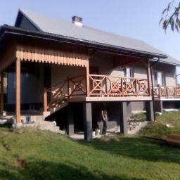 Larhome - Drewniana zabudowa balkonu z dekoracyjnymi balustradami i podbitką, widok na dom z szarą elewacją i metalowym dachem, trawnik z widocznymi śladami budowy.