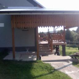 Larhome - Zadaszona weranda z drewnianą balustradą i ozdobnymi wykończeniami, widok z zewnątrz na tle zieleni, częściowo ocieniona, z widoczną osobą pod konstrukcją.