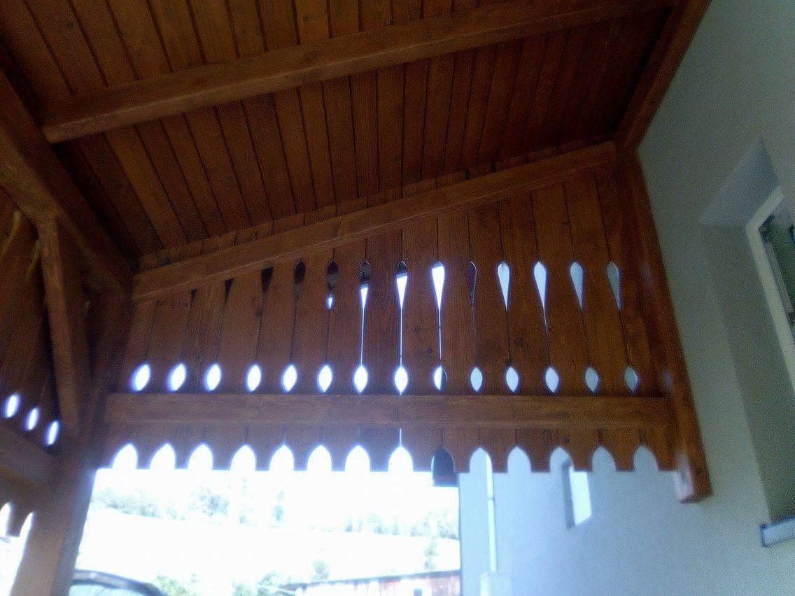 Drewniana zabudowa balkonu z dekoracyjnymi wycięciami, widok od wewnątrz, ukazujący konstrukcję dachu i fragment ściany budynku.