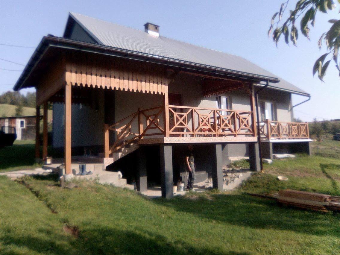 Drewniana zabudowa balkonu z dekoracyjnymi balustradami i podbitką, widok na dom z szarą elewacją i metalowym dachem, trawnik z widocznymi śladami budowy.