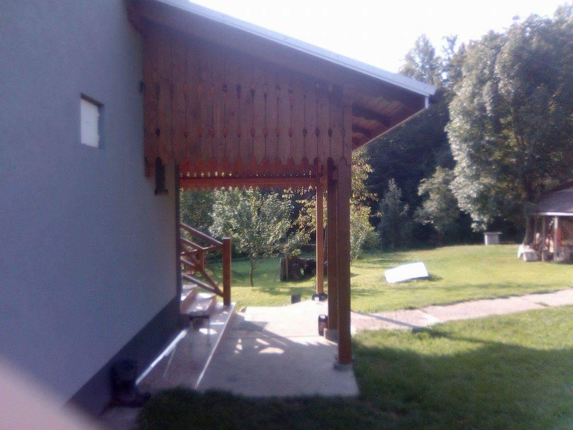 Drewniana weranda z ozdobnymi wycięciami, przylegająca do szarego budynku, widok na zielony trawnik i drzewa w tle.