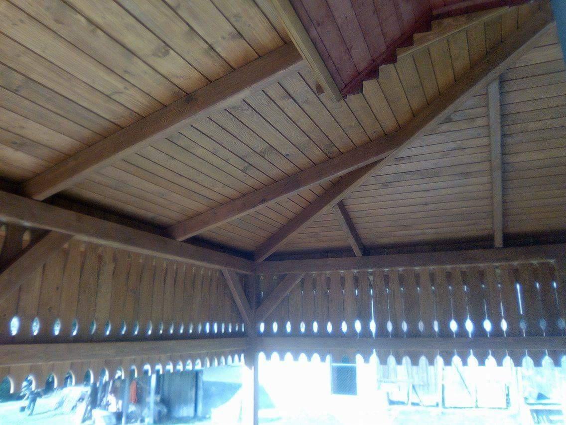 Drewniana konstrukcja zadaszenia z widocznymi belkami stropowymi i ozdobną balustradą, wykonana z jasnego drewna, widok od wewnątrz, z niewyraźnym tłem zewnętrznym.