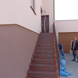 Janbud - Zewnętrzne schody wejściowe do domu, zabezpieczone taśmą malarską, z widoczną elewacją w dwóch odcieniach beżu i pracownikami w tle.
