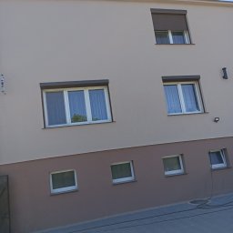 Janbud - Dwukolorowa elewacja budynku z oknami, brązowe rolety zewnętrzne na jednym oknie, widoczne przewody na chodniku.