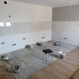 Janbud - Wnętrze w trakcie prac wykończeniowych: położone płytki podłogowe i częściowo ścienne imitujące marmur, widoczne puszki elektryczne, narzędzia, krzesło i wiaderka po farbie.