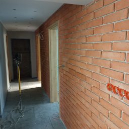 Janbud - Wnętrze korytarza z wykończoną ścianą z cegły klinkierowej, widoczne puszki elektryczne, szary gres na podłodze i lampa budowlana. Świeżo otynkowane ściany po lewej stronie.