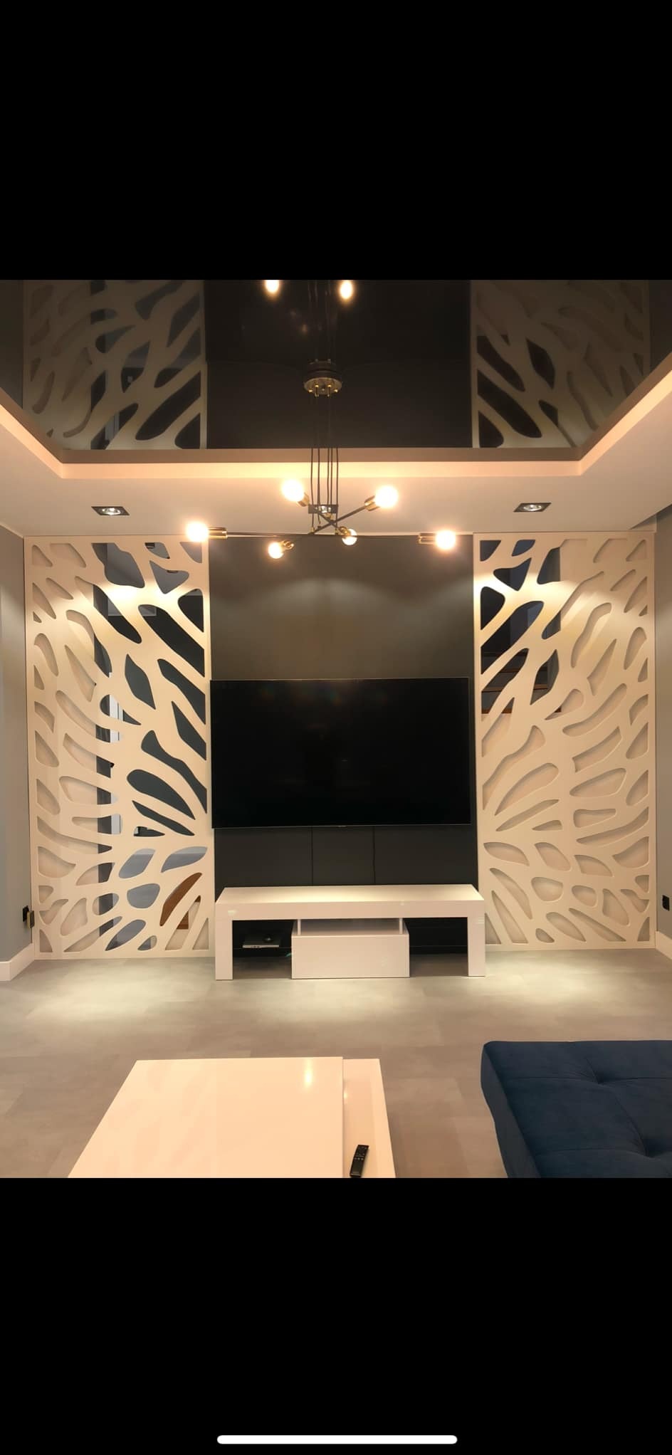 Nowoczesny salon z ażurowymi panelami dekoracyjnymi po bokach telewizora, podwieszany sufit z oświetleniem punktowym i designerska lampa wisząca