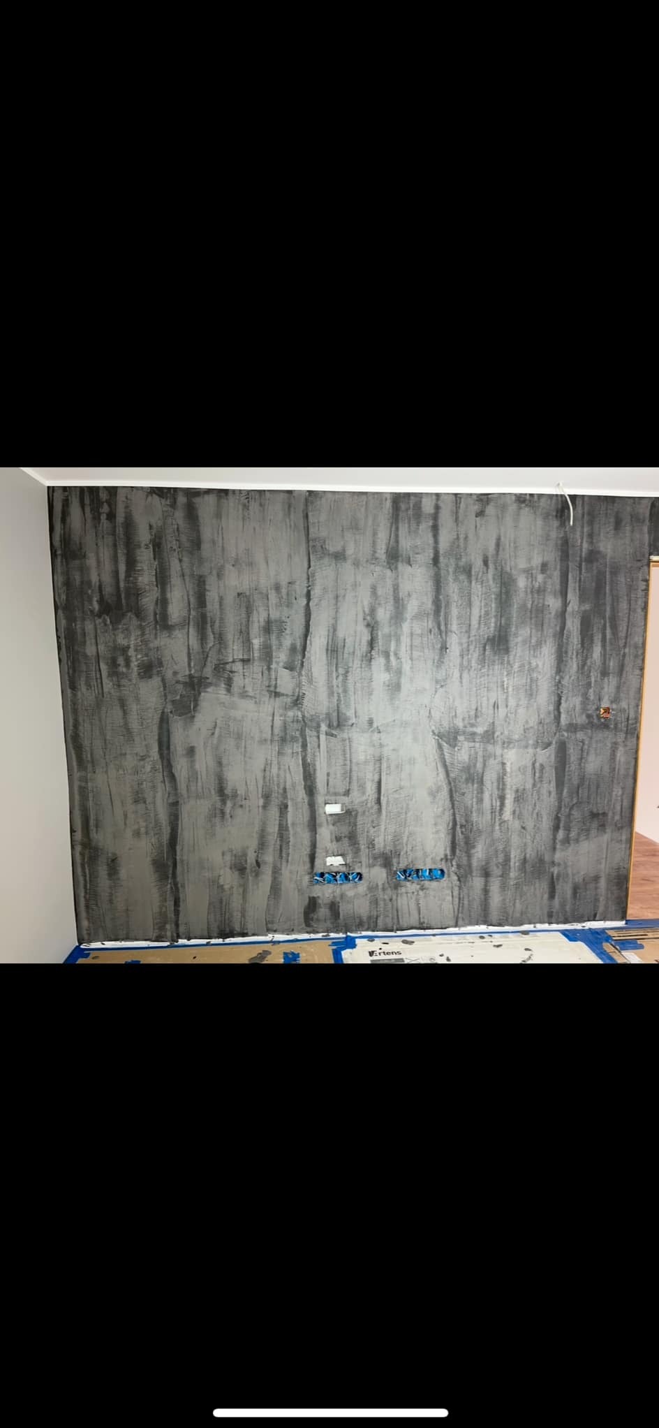 Ściana z efektem dekoracyjnym betonu, zabezpieczona taśmą malarską i kartonem na podłodze, widoczne puszki elektryczne.