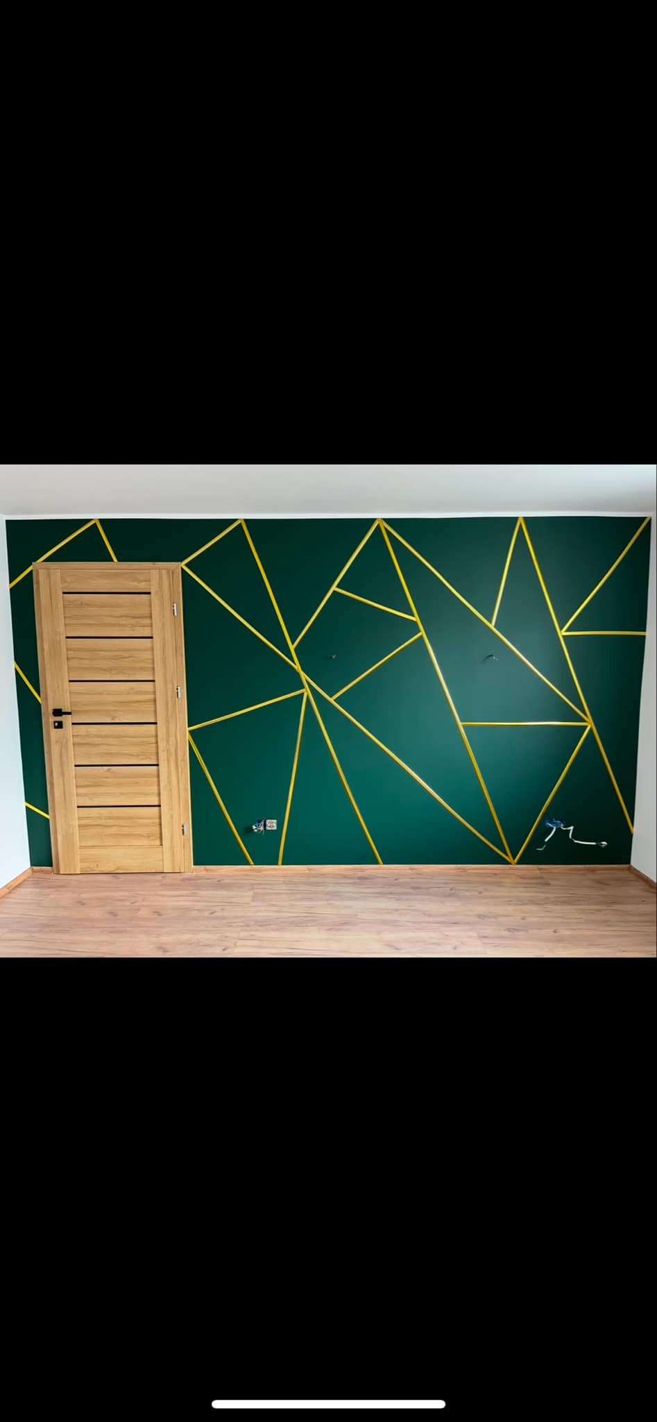 Ściana pomalowana na ciemnozielono z geometrycznym wzorem z żółtych listew, drewniane panele podłogowe i drzwi z naturalnego drewna.