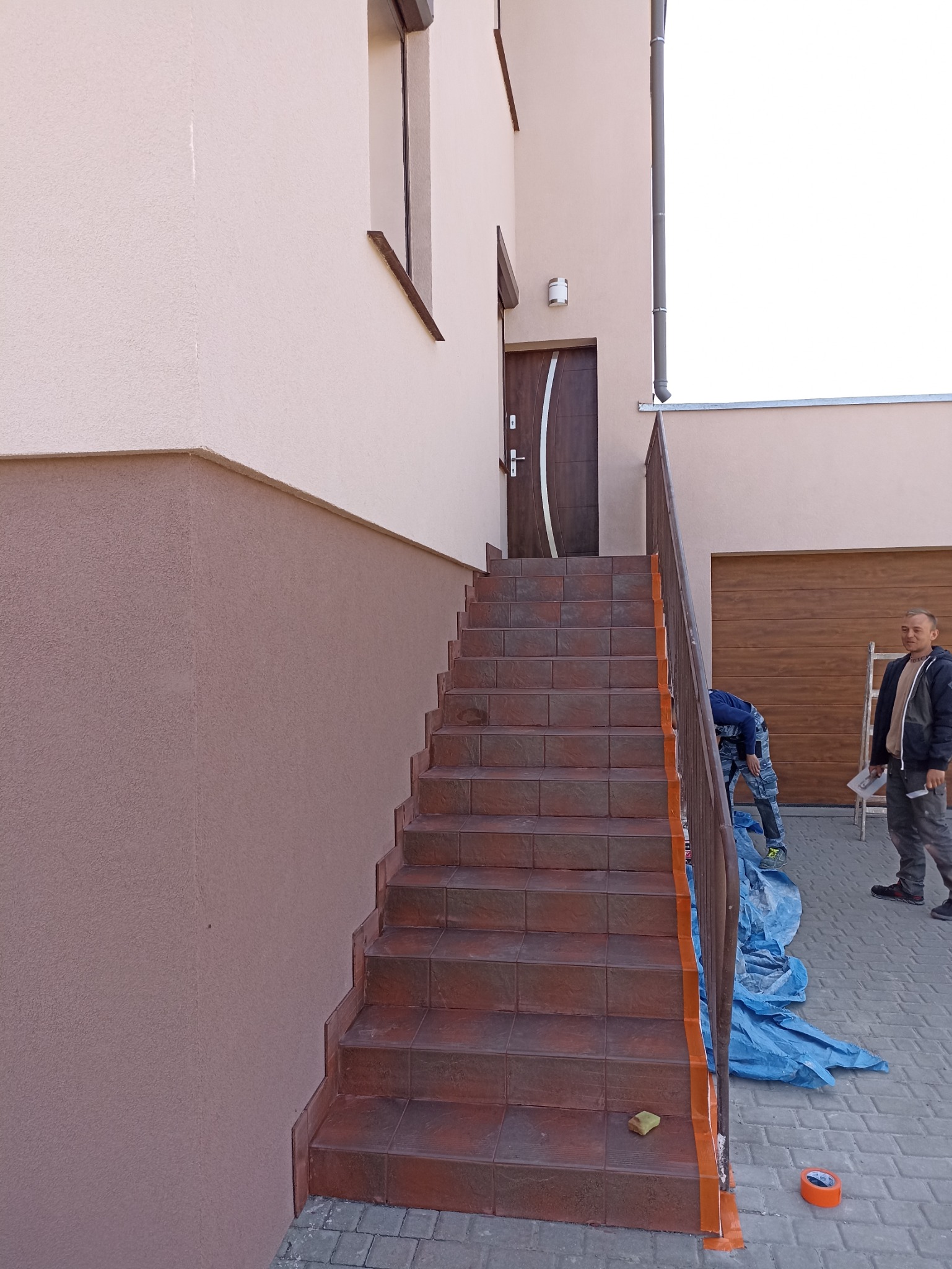 Zewnętrzne schody wejściowe do domu, zabezpieczone taśmą malarską, z widoczną elewacją w dwóch odcieniach beżu i pracownikami w tle.
