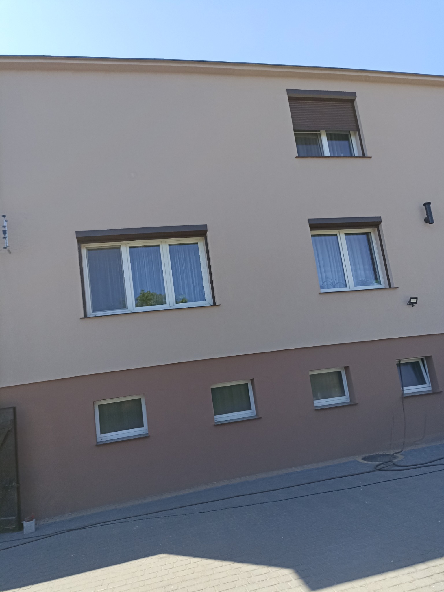 Dwukolorowa elewacja budynku z oknami, brązowe rolety zewnętrzne na jednym oknie, widoczne przewody na chodniku.