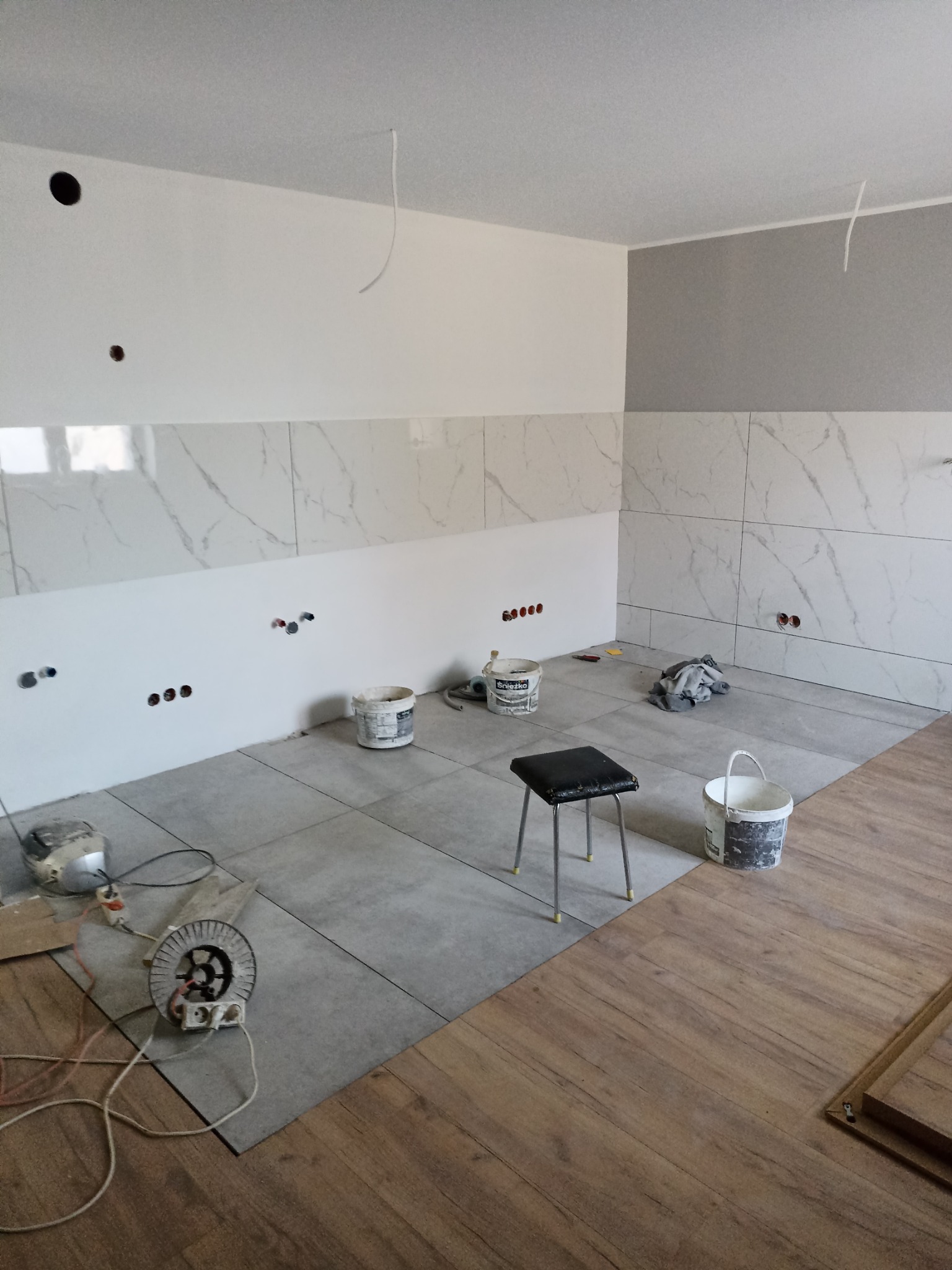 Wnętrze w trakcie prac wykończeniowych: położone płytki podłogowe i częściowo ścienne imitujące marmur, widoczne puszki elektryczne, narzędzia, krzesło i wiaderka po farbie.
