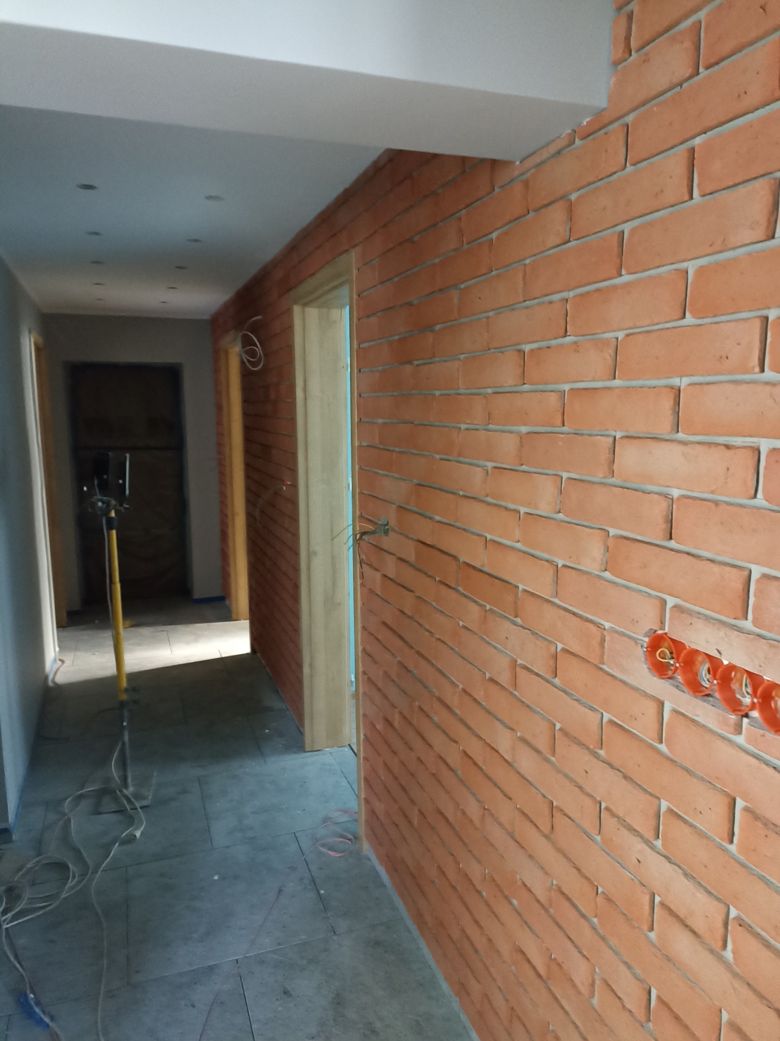 Wnętrze korytarza z wykończoną ścianą z cegły klinkierowej, widoczne puszki elektryczne, szary gres na podłodze i lampa budowlana. Świeżo otynkowane ściany po lewej stronie.