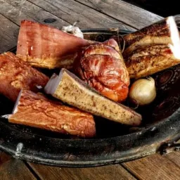 Wędliny i czosnek ułożone w metalowej misie na drewnianym stole, obok gliniany kubek.