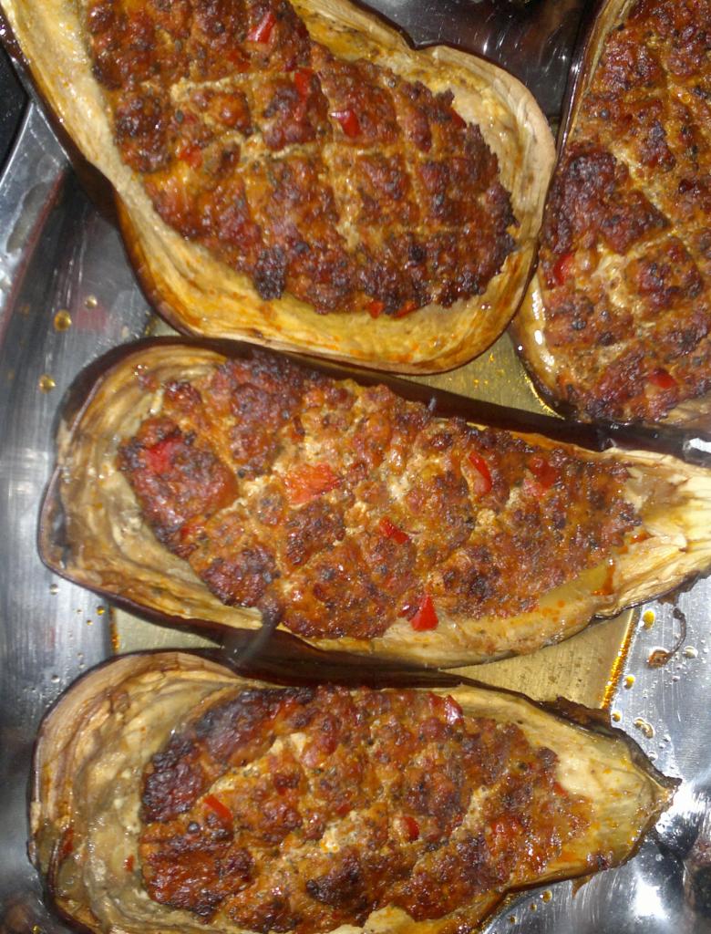 Cztery połówki pieczonych bakłażanów wypełnionych farszem mięsnym z czerwoną papryką, ułożone na metalowej tacy.