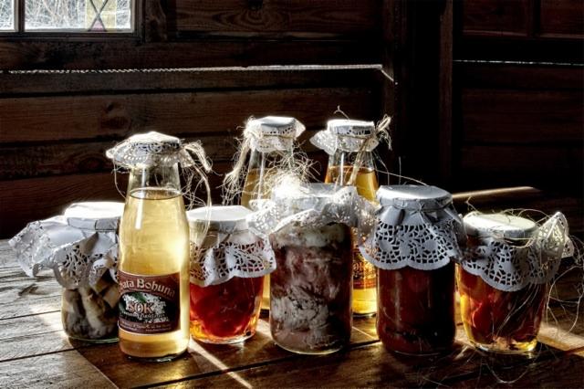 Słoiki i butelki z domowymi przetworami, zakryte serwetkami, ustawione na drewnianym stole na tle drewnianej ściany i okna.