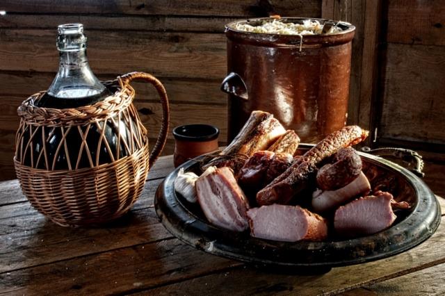 Rustykalna kompozycja z wędlinami, boczkiem, czosnkiem, kapustą w glinianym garnku i butelką wina w wiklinowym koszu na drewnianym stole.