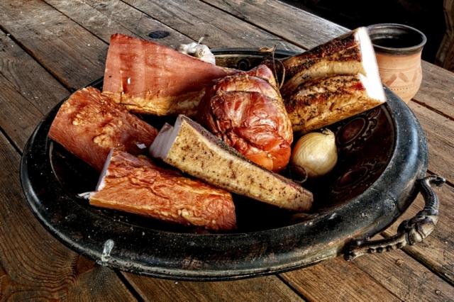 Wędliny i czosnek ułożone w metalowej misie na drewnianym stole, obok gliniany kubek.