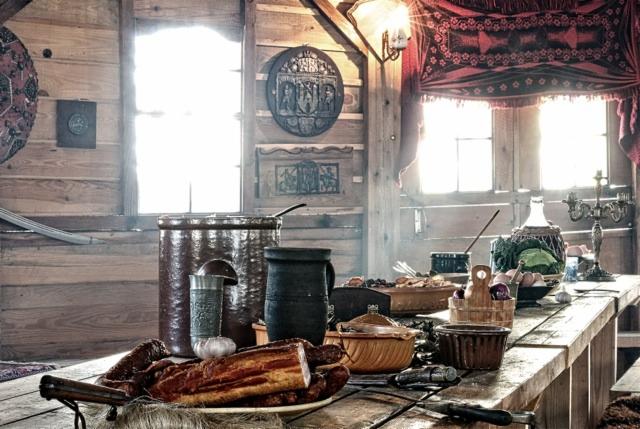 Rustykalny stół w drewnianej chacie z wędlinami, naczyniami ceramicznymi i metalowymi, dekoracjami ściennymi i oknami z czerwonymi zasłonami.