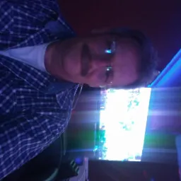 Mężczyzna w okularach i koszuli w kratę, oświetlony niebieskim światłem, patrzy w ekran. Zdjęcie z bliska, wykonane w pomieszczeniu z widocznym oświetleniem LED.