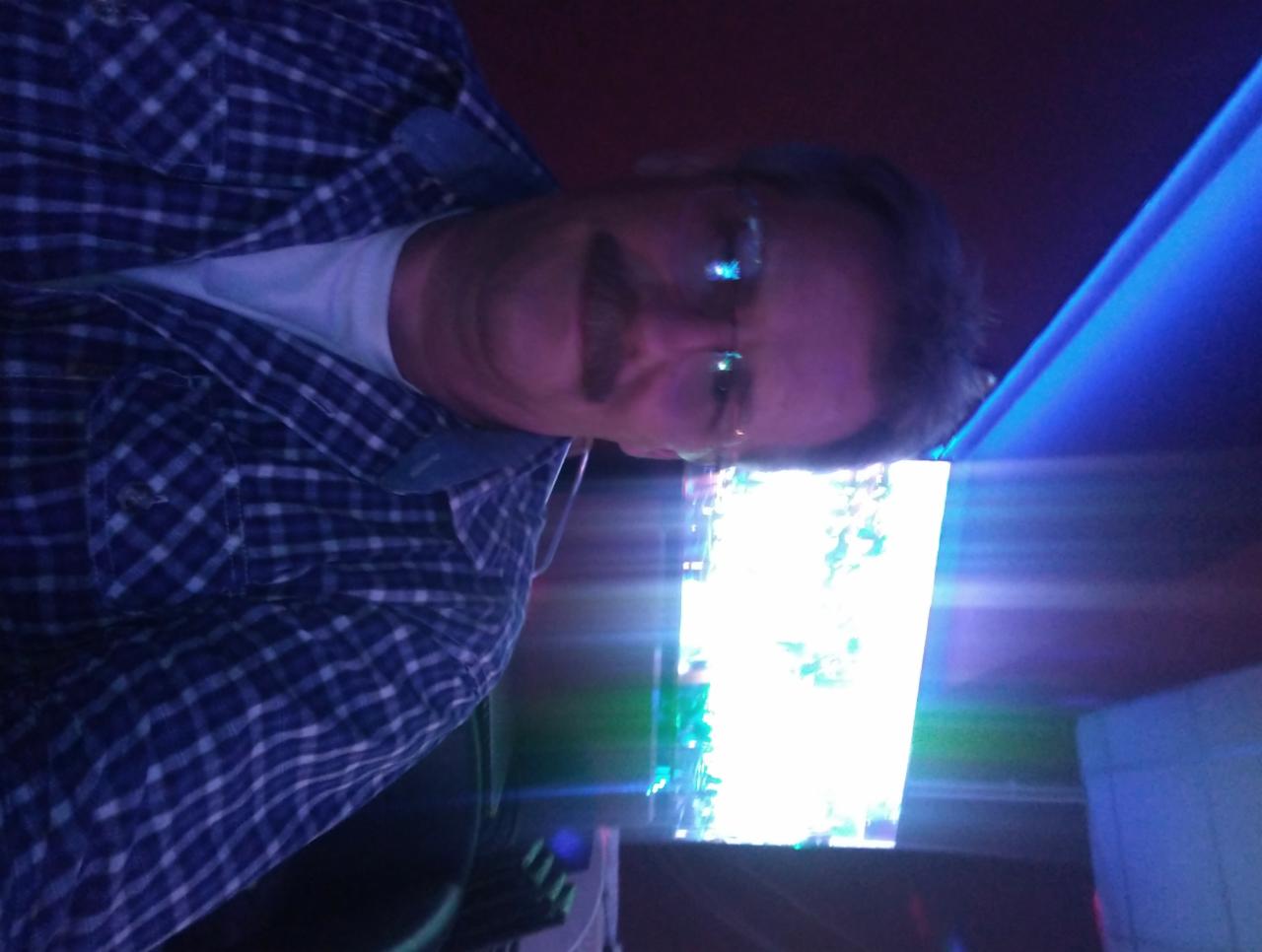 Mężczyzna w okularach i koszuli w kratę, oświetlony niebieskim światłem, patrzy w ekran. Zdjęcie z bliska, wykonane w pomieszczeniu z widocznym oświetleniem LED.