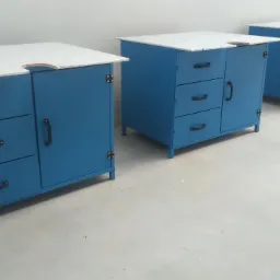 Trzy niebieskie szafki warsztatowe z szufladami i blatem z białej płyty, ustawione w rzędzie w jasnym pomieszczeniu.