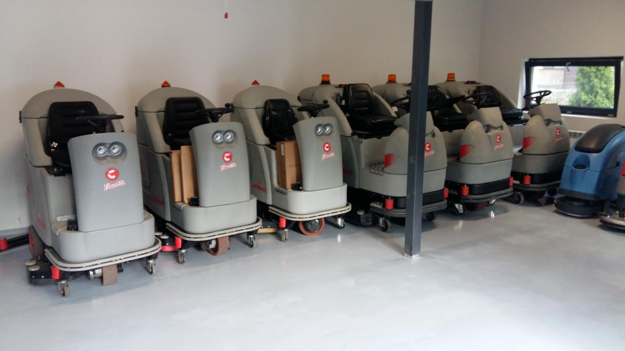 Rząd szarych, samojezdnych maszyn czyszczących podłogi, ustawionych w rzędzie w jasnym pomieszczeniu magazynowym z oknem w tle.