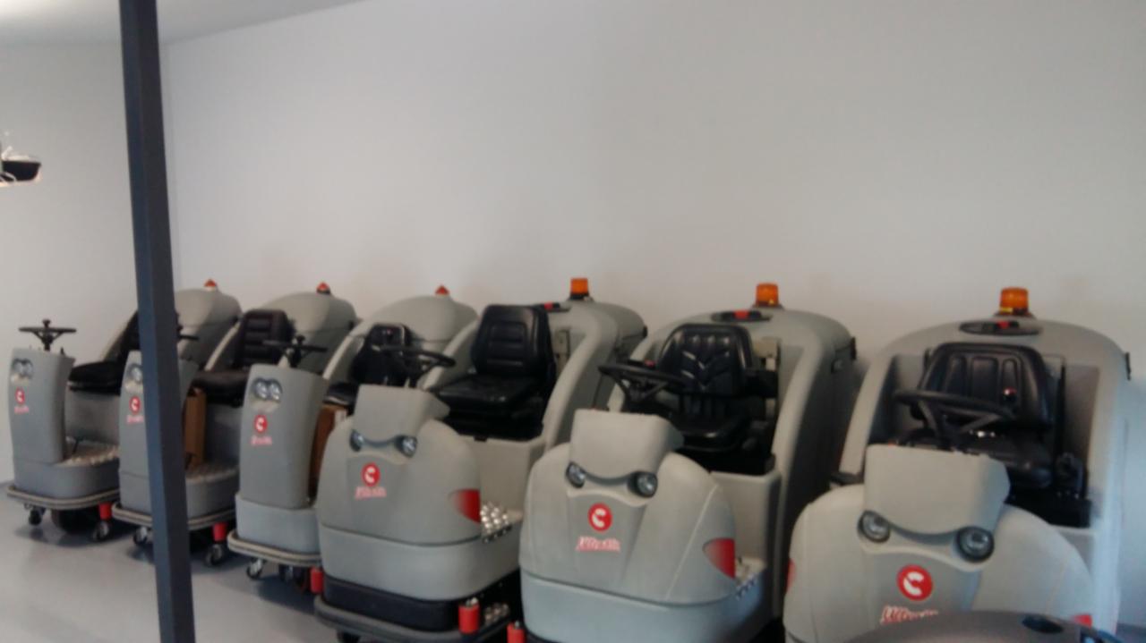 Rząd szarych, samojezdnych maszyn czyszczących podłogi z czarnymi siedzeniami i pomarańczowymi światłami ostrzegawczymi, ustawionych w jasnym pomieszczeniu.