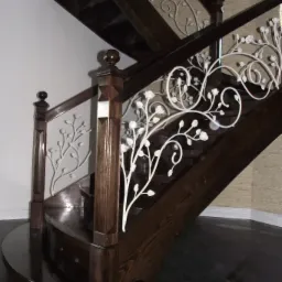 Ciemne drewniane schody z ozdobną, białą, metalową balustradą z motywem kwiatowym, widoczne od dołu, z fragmentem ściany w tle.