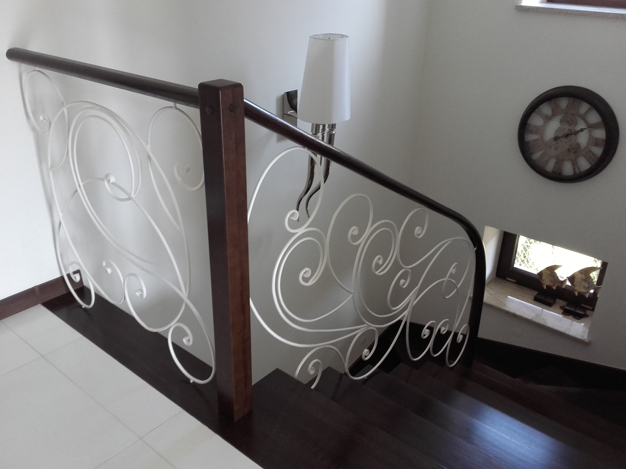 Białe, ozdobne metalowe balustrady z drewnianymi słupkami na ciemnych drewnianych schodach, widok z góry, zegar na ścianie w tle.