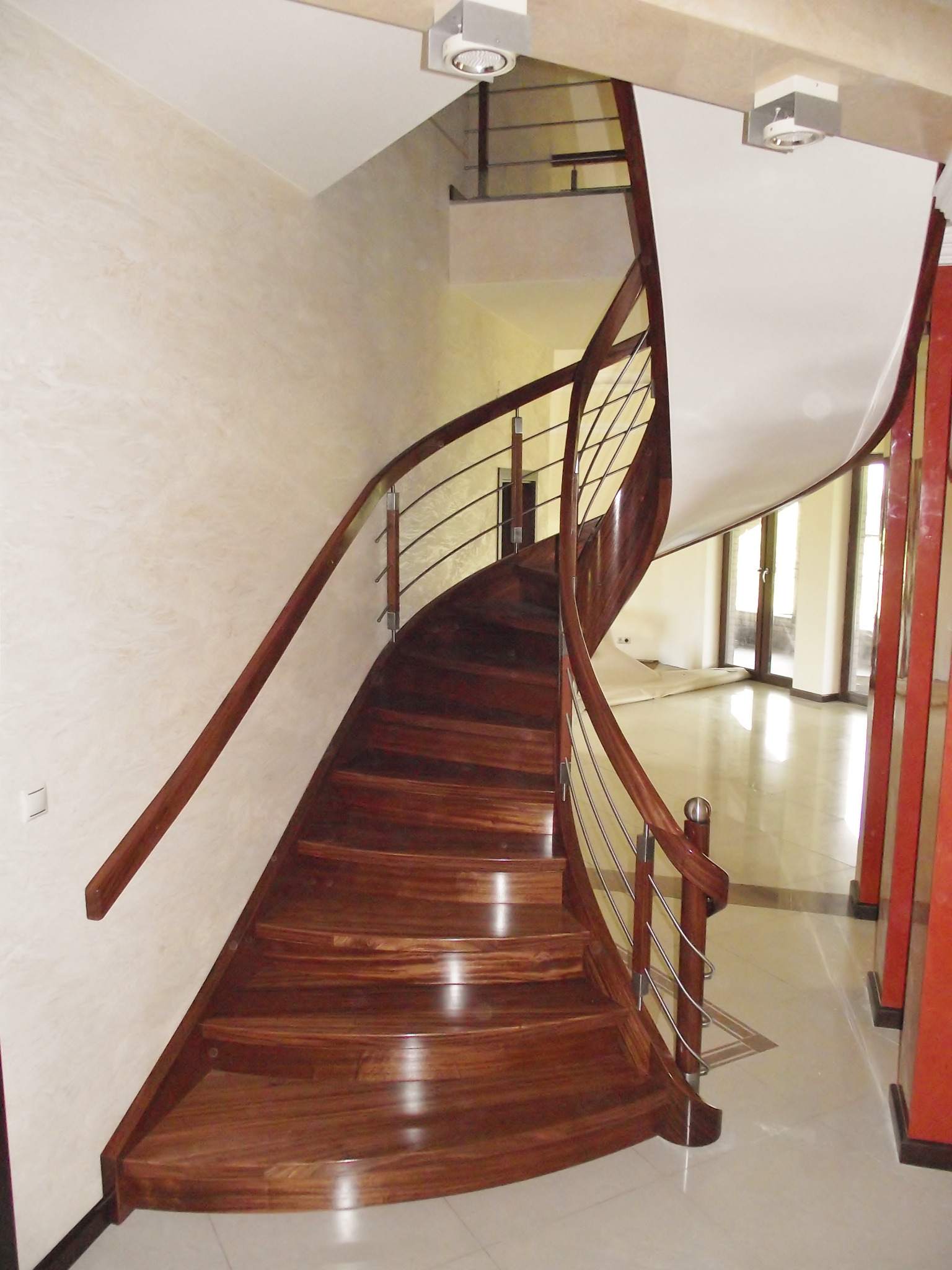 Kręte schody z ciemnego drewna z metalową balustradą w jasnym wnętrzu, widok z dołu na górę, z widocznymi lampami sufitowymi i fragmentem podłogi z jasnych płytek.