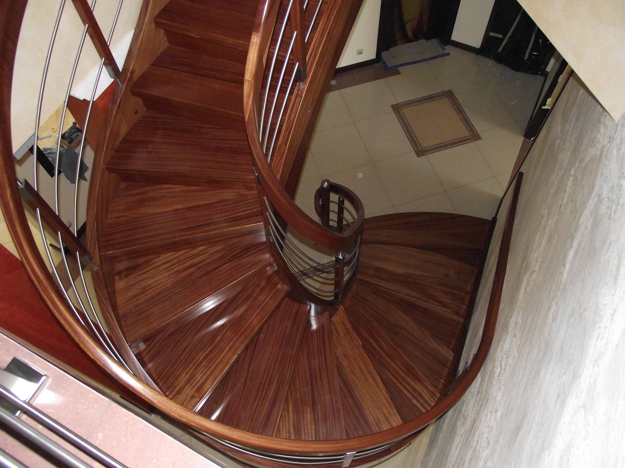 Wysoki kąt widzenia na spiralne schody z ciemnego drewna z metalowymi balustradami, widoczny fragment podłogi wyłożonej jasnymi płytkami.