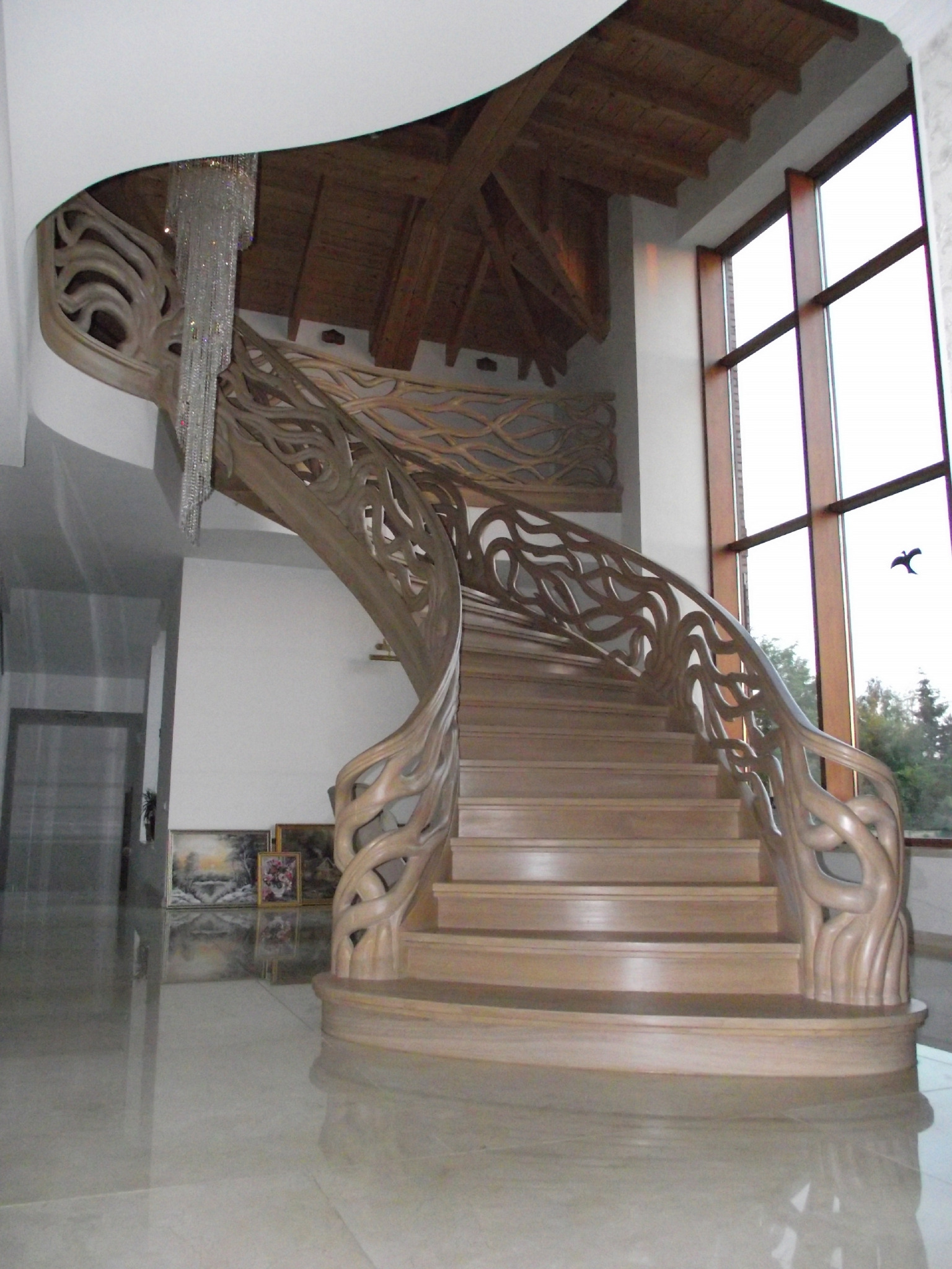 Spiralne schody drewniane z ozdobnymi, ażurowymi balustradami w Drużbicach, oświetlone kryształowym żyrandolem i dużym oknem z widokiem na zieleń.