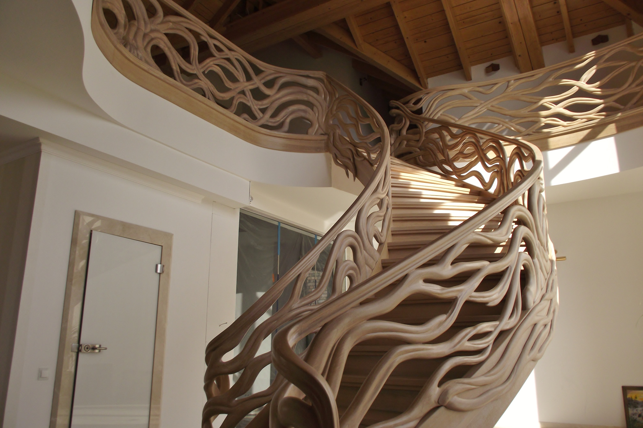 Kręte, drewniane schody z rzeźbioną balustradą przypominającą korzenie drzew, prowadzące na górę w jasnym wnętrzu z drewnianym sufitem.