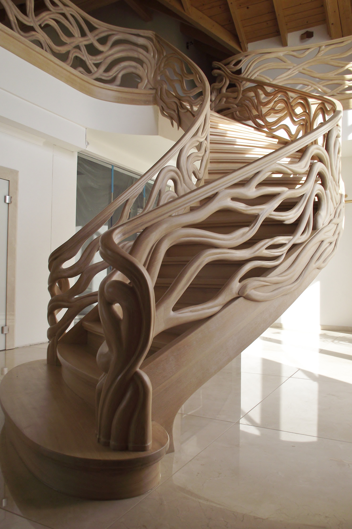 Spiralne schody z jasnego drewna z rzeźbioną balustradą przypominającą gałęzie drzew, widok od dołu na tle białych ścian i błyszczącej podłogi.
