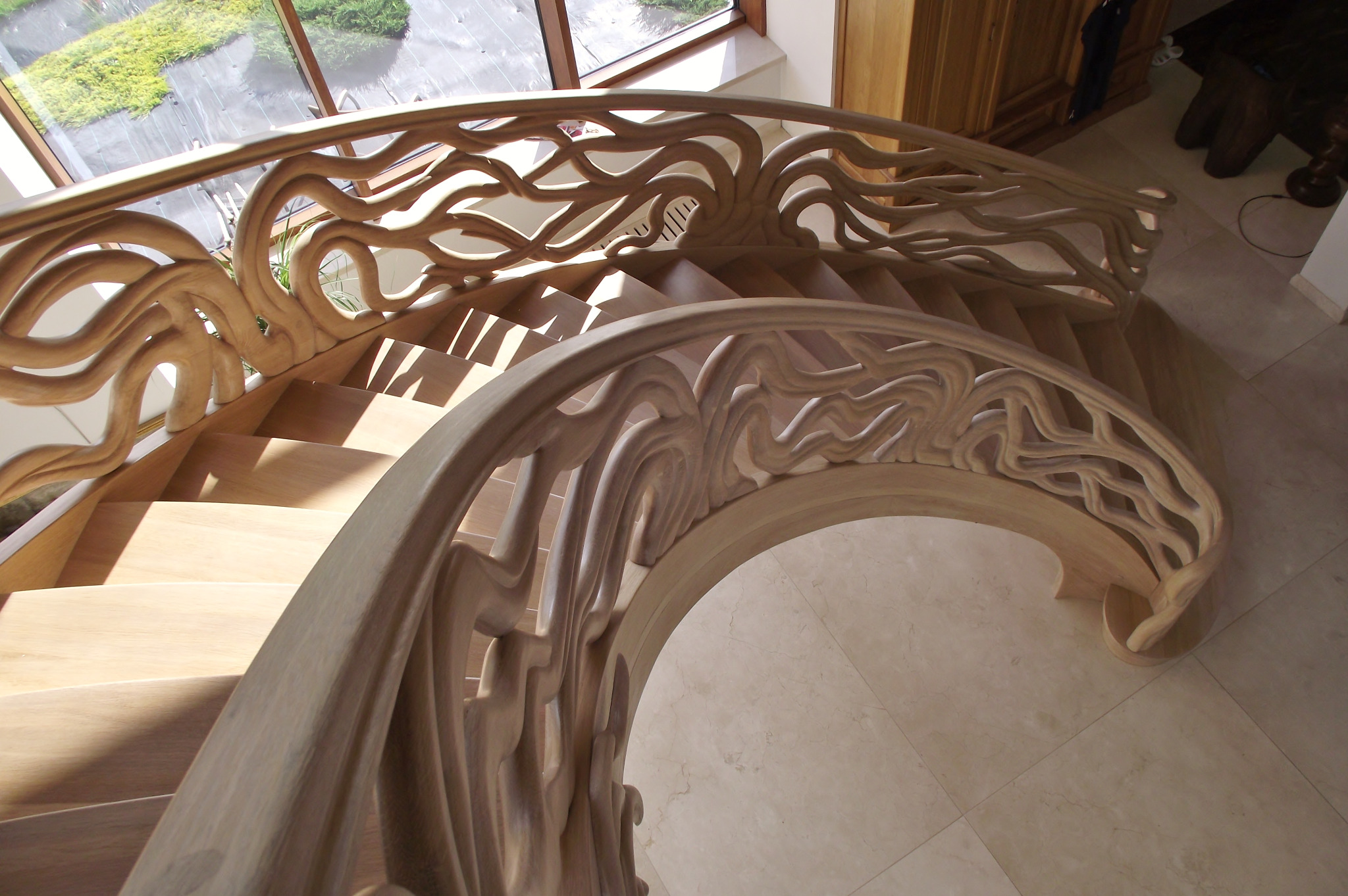 Kręte, jasne drewniane schody z ażurową balustradą w kształcie fal, widok z góry, naturalne światło z okna.