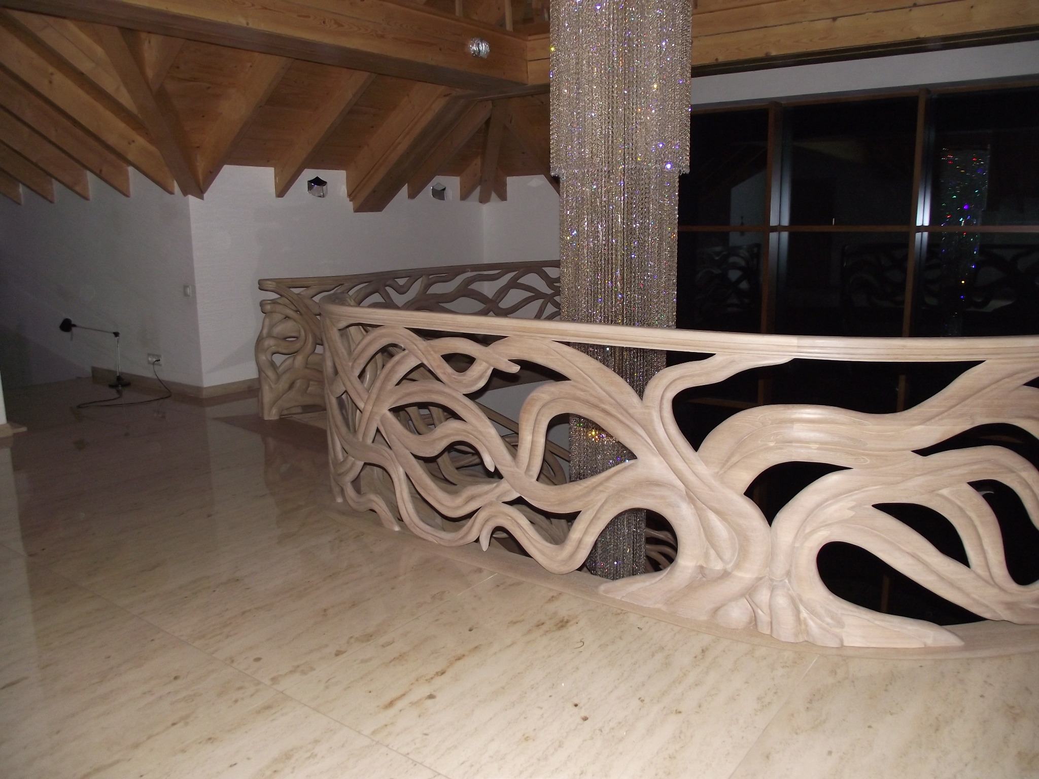 Ażurowa, jasna balustrada schodów z motywem organicznym, drewniany strop z widocznymi belkami, kryształowy żyrandol w nowoczesnym wnętrzu.