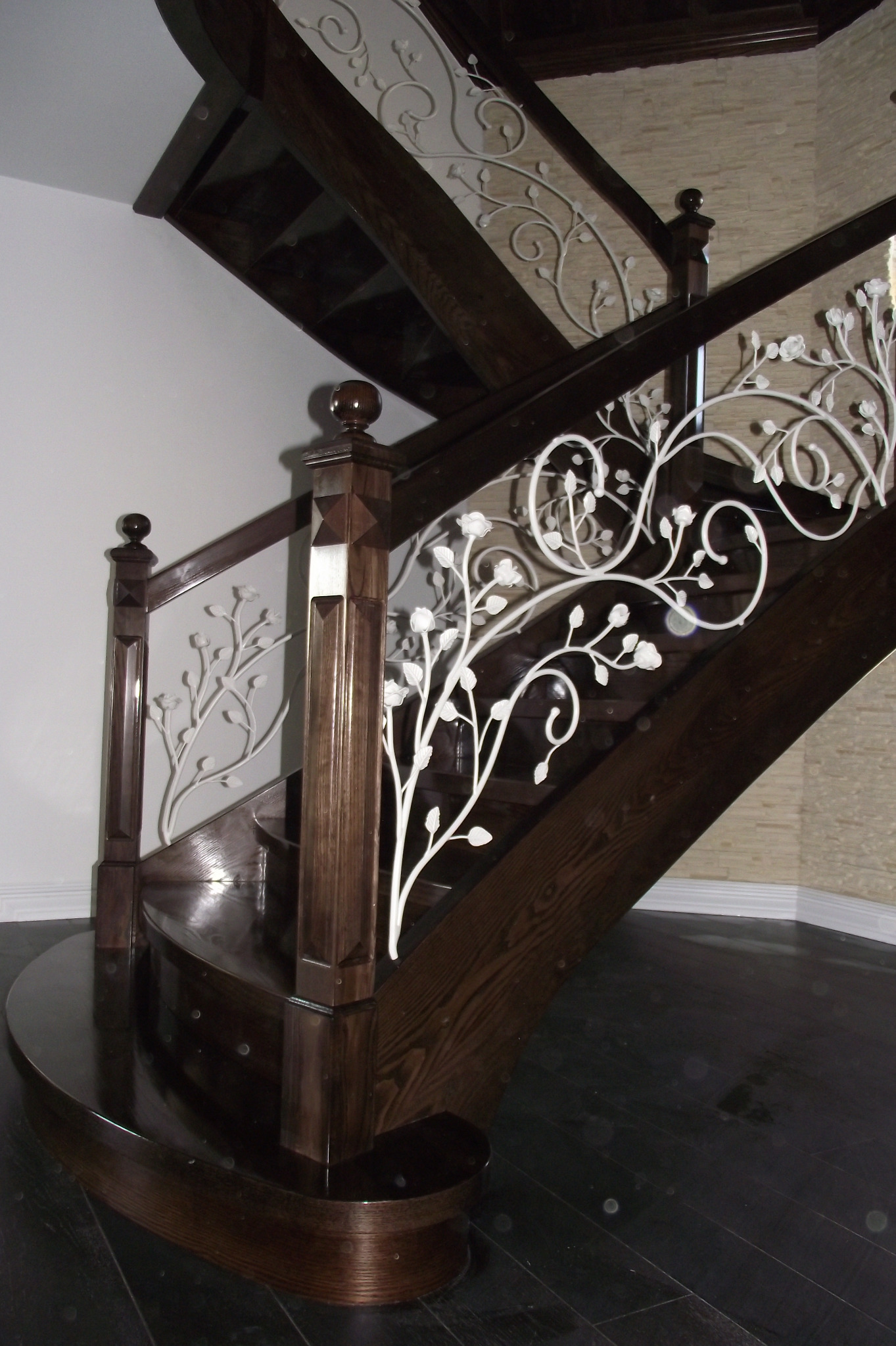 Ciemne drewniane schody z ozdobną, białą, metalową balustradą z motywem kwiatowym, widoczne od dołu, z fragmentem ściany w tle.