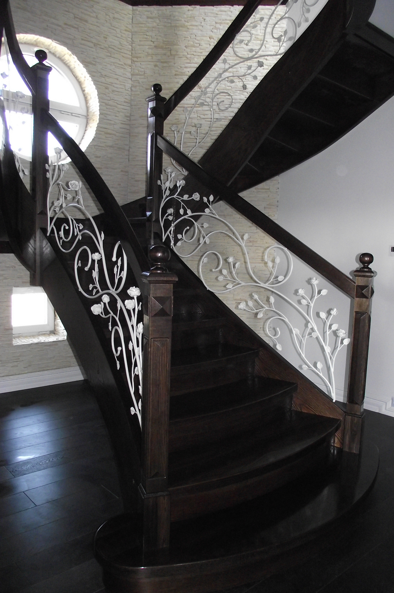 Ciemne drewniane schody z białą, metalową balustradą zdobioną motywem roślinnym, widok od dołu z elementami okrągłego okna i kamiennej ściany w tle.